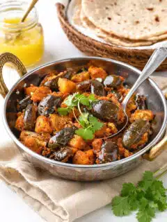 Eggplant Aloo in a small kadai with baby eggplants, potatoes, fresh cilantro, chapatis, and ghee in the background.