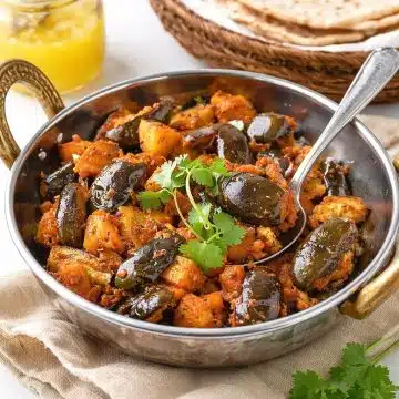 Eggplant Aloo in a small kadai with baby eggplants, potatoes, fresh cilantro, chapatis, and ghee in the background.