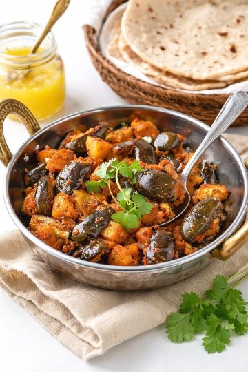 Eggplant Aloo in a small kadai with baby eggplants, potatoes, fresh cilantro, chapatis, and ghee in the background.