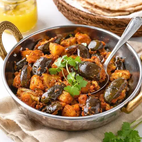 Eggplant Aloo in a small kadai with baby eggplants, potatoes, fresh cilantro, chapatis, and ghee in the background.