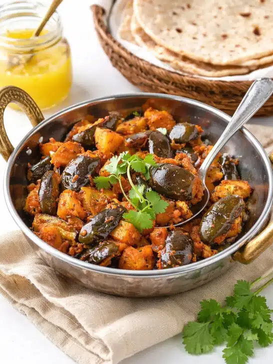 Eggplant Aloo in a small kadai with baby eggplants, potatoes, fresh cilantro, chapatis, and ghee in the background.