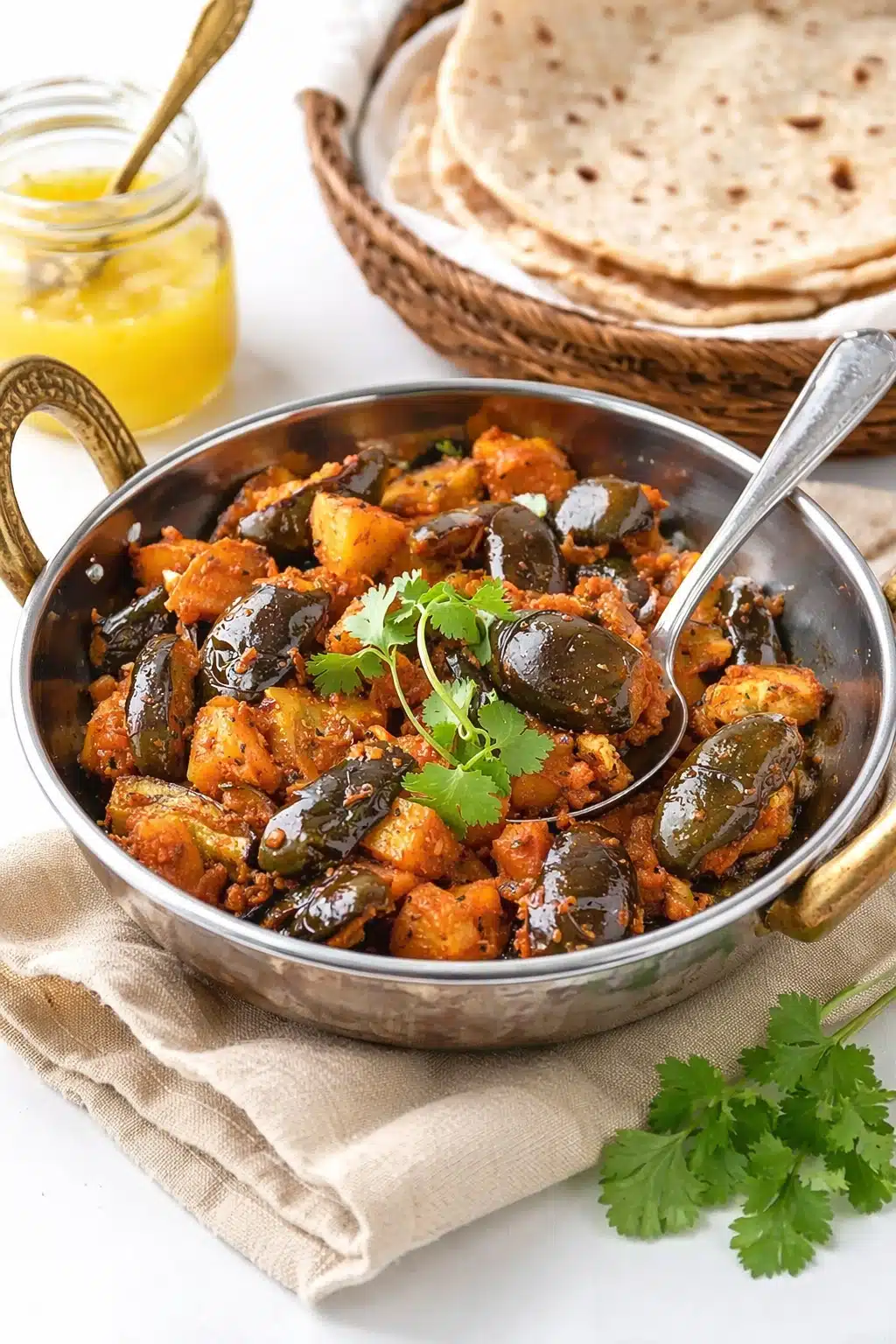 Achari Baingan Aloo in a small kadai with baby eggplants, potatoes, fresh cilantro, chapatis, and ghee in the background.