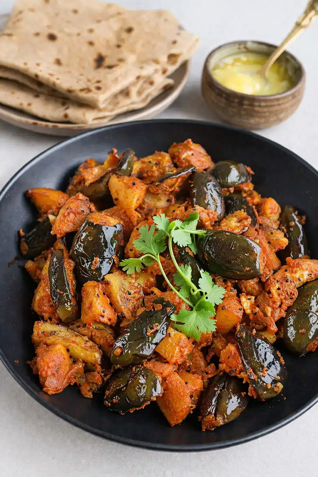 Tangy and spicy Achari Aloo served in a flat black bowl with Indian flatbread and ghee.