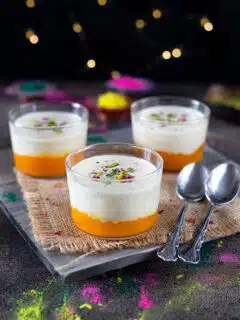 Three glasses of layered dessert with a mango base and creamy topping, served on a rustic wooden board with a blurred background of twinkling lights.
